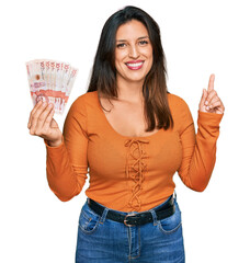 Beautiful hispanic woman holding 10 colombian pesos banknotes surprised with an idea or question pointing finger with happy face, number one
