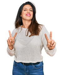 Beautiful hispanic woman wearing casual sweater smiling looking to the camera showing fingers doing...