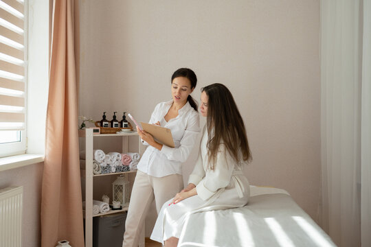 Confident focused massage therapist in uniform holding clipboard and consulting female customer sitting on bed about therapy procedure in salon