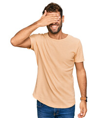 Handsome young man with beard wearing casual tshirt smiling and laughing with hand on face covering eyes for surprise. blind concept.