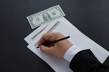 A businessman's hand in a suit draws five dollars with a pencil on a white piece of paper, and one dollar lies next to it. Tax fraud, financial crime