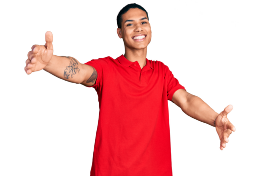 Young hispanic man wearing casual red t shirt looking at the camera smiling with open arms for hug. cheerful expression embracing happiness.