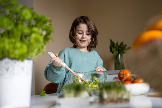 Elementary cute girl wearing casuals mixing vegetables with spatula in glass bowl while preparing salad at home