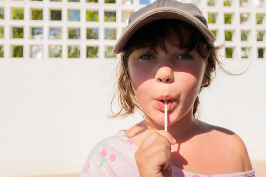 Closeup portrait of cute serious girl in casual clothes and cap eating sweet lollipop and staring at camera while standing on the street