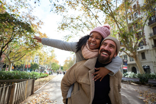 Portrait of young cheerful multiracial happy couple in love hugging and smiling in piggyback pointing somewhere outdoors at street. Smiling people dressed in winter clothes having fun together