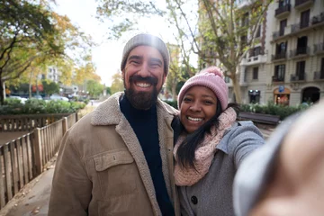 Foto op Aluminium Winter portrait of happy young tourist couple taking selfie outdoors looking at camera in a city. Smiling multi-ethnic people traveling together. Cheerful man and woman posing for photo with phone. © CarlosBarquero