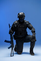 Fototapeta premium Soldier in black military uniform for city with helmet, action camera and machine gun, photo in studio. Army man stands on one knee, defensive position