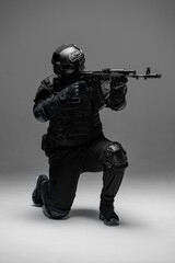 Soldier in black military uniform for city with helmet, action camera and machine gun, photo in studio. Army man stands on one knee, defensive position
