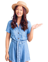 Young beautiful chinese girl wearing summer hat smiling cheerful presenting and pointing with palm of hand looking at the camera.