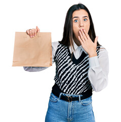 Young brunette teenager holding take away paper bag covering mouth with hand, shocked and afraid for mistake. surprised expression