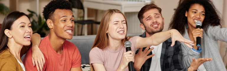 Obraz premium Karaoke time. Group of cheerful young multicultural friends playing karaoke at home, singing with microphone, having great time together while sitting on the couch in the living room. Karaoke party