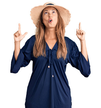 Middle age hispanic woman wearing summer hat amazed and surprised looking up and pointing with fingers and raised arms.