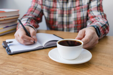 unrecognizable man holding cup of coffee and writing in notepad on wooden table, studying or working, sitting at the desk with stack of books, focus on foreground