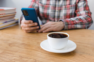 Cup of coffee on wooden table, unrecognizable man using smartphone, sitting at the desk with stack of books, focus on foreground