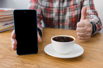 Unrecognizable man showing smartphone screen and big thumbs up, sitting at the wooden table with coffee cup and stack of books