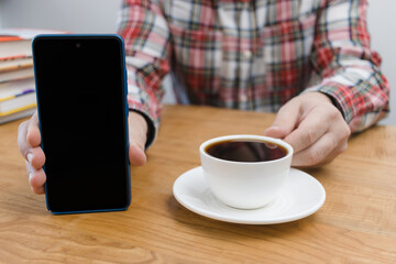 Fototapeta na wymiar Unrecognizable man showing smartphone screen, sitting at the wooden table with coffee cup and stack of books