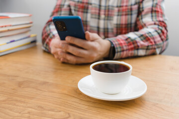 Cup of coffee on wooden table, unrecognizable man using smartphone, sitting at the desk with stack of books, focus on foreground