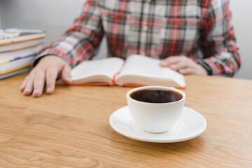 Cup of coffee on wooden table, unrecognizable man reading book, studying or working, sitting at the desk with stack of books, focus on foreground