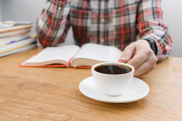 Unrecognizable man reading book and holding cup of coffee, studying or working, sitting at the table with stack of books, focus on foreground