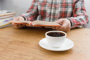 Cup of coffee on wooden table, unrecognizable man reading book, studying or working, sitting at the desk with stack of books, focus on foreground