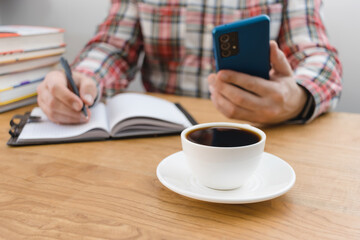 Fototapeta na wymiar Cup of coffee on wooden table, unrecognizable man writing in notepad and using smartphone, studying or working, sitting at the desk with stack of books, focus on foreground