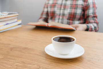 Cup of coffee on wooden table, unrecognizable man reading book, studying or working, sitting at the desk with stack of books, focus on foreground