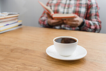 Cup of coffee on wooden table, unrecognizable man reading book, studying or working, sitting at the desk with stack of books, focus on foreground
