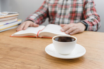 Cup of coffee on wooden table, unrecognizable man reading book, studying or working, sitting at the desk with stack of books, focus on foreground