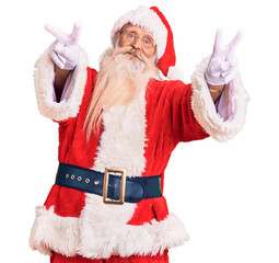 Old senior man with grey hair and long beard wearing traditional santa claus costume smiling with tongue out showing fingers of both hands doing victory sign. number two.