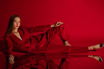  Fashionable confident woman wearing trendy red suit blazer, classic trousers, metallic silver color shoes, laying, posing on red background. Full-length studio fashion portrait. Copy, empty space 