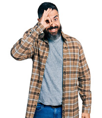 Hispanic man with beard wearing casual shirt doing ok gesture with hand smiling, eye looking through fingers with happy face.