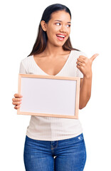 Young beautiful latin girl holding empty white chalkboard pointing thumb up to the side smiling happy with open mouth