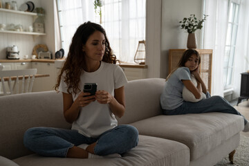 Caring puzzled mother sits on couch with mobile phone and looks back at offended teenage daughter after punishment. Young reserved female in casual clothes wants to make call to child psychologist