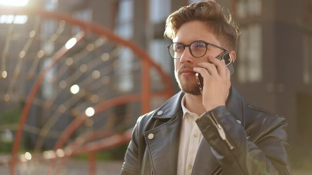 Close-up of a young, stylish man in glasses answering a phone call against the backdrop of a modern building