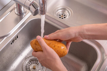 Female hands washing potato with hand under running water in sink in the kitchen. Vegetables,...