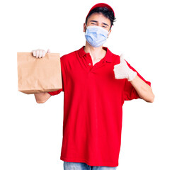 Young hispanic man wearing delivery uniform and medical mask holding paper bag smiling happy and positive, thumb up doing excellent and approval sign
