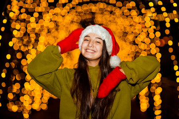 A young, beautiful girl walks in a Santa hat, in the evening city, decorated with New Year's lights
