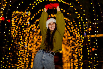 A young, beautiful girl walks in a Santa hat, in the evening city, decorated with New Year's lights