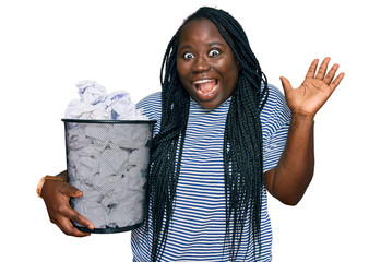 Young black woman with braids holding paper bin full of crumpled papers celebrating victory with...