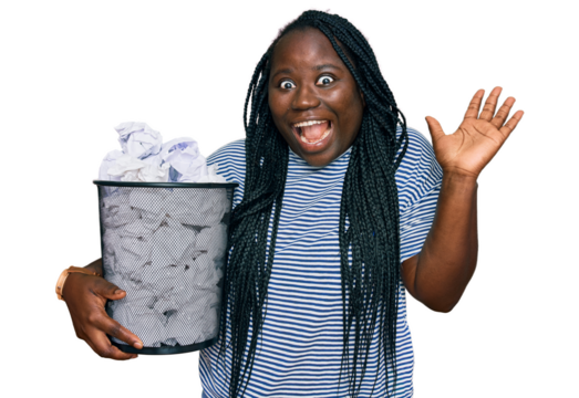 Young black woman with braids holding paper bin full of crumpled papers celebrating victory with happy smile and winner expression with raised hands