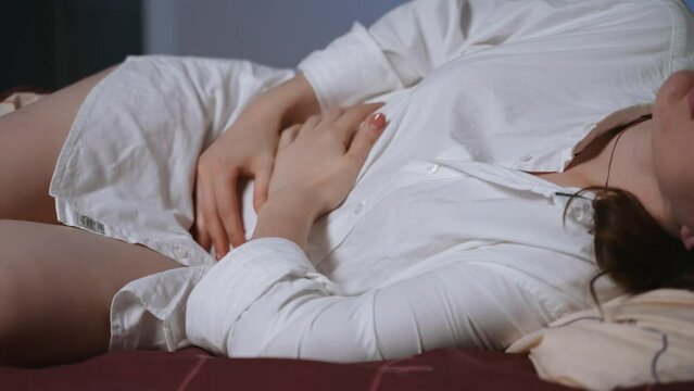 A young woman lying on her side on the bed writhes from stomach pain. Dressed in a white shirt, she is in a state of upset or discomfort. The bed is covered with a brown patterned sheet. 