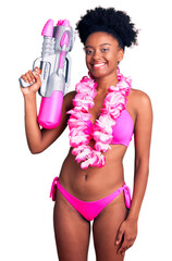 Young african american woman wearing bikini and hawaiian lei holding water gun looking positive and...