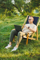 Freelancer man sits on a wooden chair in nature and works online on a laptop. A man travels and works remotely on a laptop computer. Office work in nature. Vacation