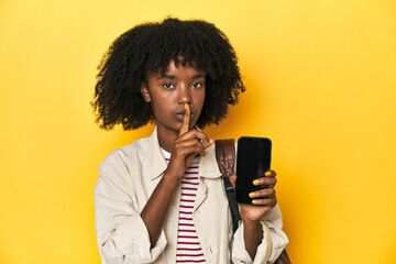 Teen girl showing smartphone, backpack on yellow background keeping a secret or asking for silence.