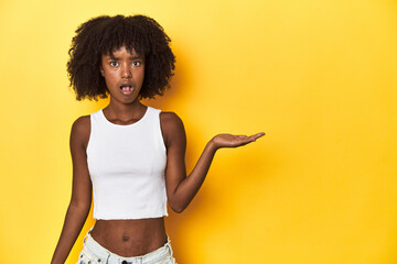 Teen girl in white tank top, yellow studio background impressed holding copy space on palm.