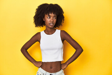 Teen girl in white tank top, yellow studio background confused, feels doubtful and unsure.