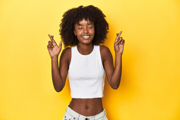 Teen girl in white tank top, yellow studio background crossing fingers for having luck