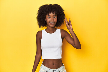 Teen girl in white tank top, yellow studio background showing a horns gesture as a revolution...