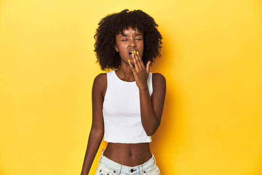 Teen girl in white tank top, yellow studio background yawning showing a tired gesture covering mouth with hand.
