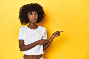 Teen girl in classic white T-shirt, yellow studio backdrop shocked pointing with index fingers to a...